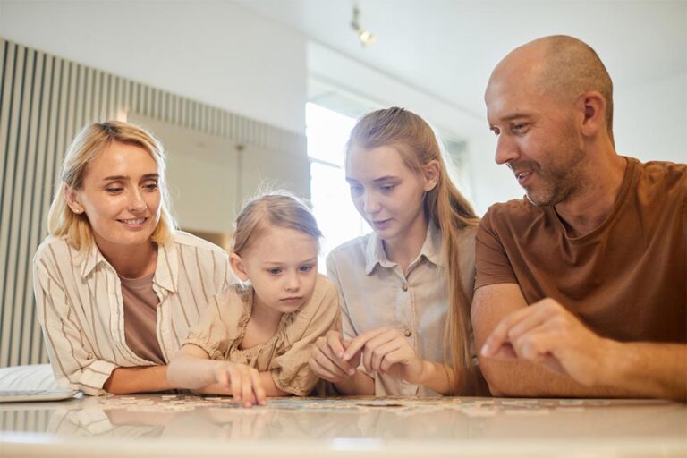 Mutter, Vater und zwei Töchter, die sich gemeinsam über einen Tisch beugen und puzzlen.