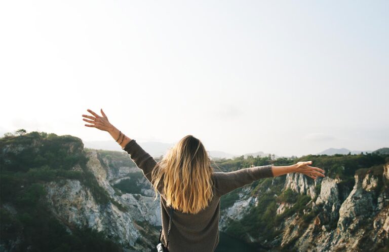 Blonde Frau, die mit ausgestreckten Armen in Richtung Horizont schaut und oben auf einem Berg steht.